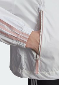 Main dans la poche latérale d'une veste blanche avec une fermeture éclair rose et des rayures roses assorties sur le poignet élastique.