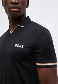 Κοντινό πλάνο ενός ατόμου που φοράει ένα μαύρο πόλο BOSS με λευκή και μπεζ επένδυση στο γιακά και το μανίκι, πάνω σε ομοιόμορφο ανοιχτό φόντο.