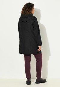 Chaqueta negra impermeable con capucha, de corte recto y bolsillos laterales, combinada con pantalones slim-fit burdeos y botas negras de tobillo.