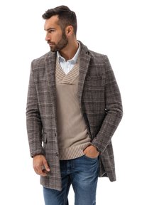 Ruskea ruudullinen bleiseri, jonka istuvuus on tarkka, villasekoituskangas, yksittäinen napitus, beige neulepaita ja vaaleansiniset farkut alla.