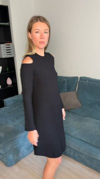 Femme en robe noire à épaules dénudées, debout près d’un canapé bleu sarcelle dans une pièce aux murs clairs et une étagère noire.