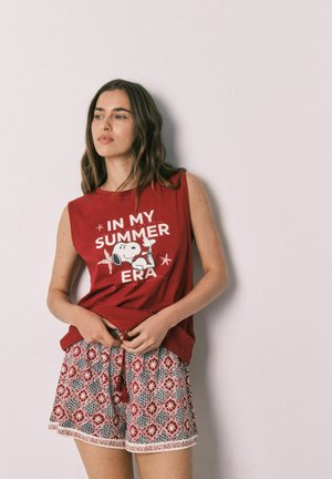 Junge Frau trägt ein rotes ärmelloses Oberteil mit Snoopy und Schriftzug, kombiniert mit rot-weiß gemusterten Shorts, steht vor einer hellen Wand.
