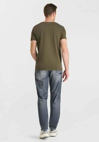 Homme se tenant de dos, portant un t-shirt vert olive, un jean bleu délavé et des baskets blanches sur un fond clair uni.