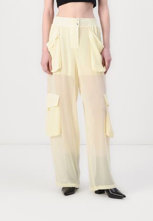 Light yellow sheer cargo pants with oversized pockets, elastic waistband, and subtle sheen; paired with black pointed heels.