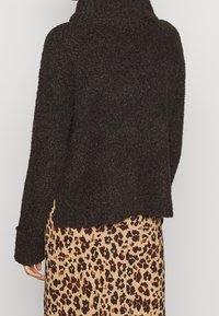 Dunkelbrauner, strukturierten Pullover mit hohem Kragen und legerem Schnitt; über einem beigefarbenen Rock mit einem schwarzen Leopardenmuster getragen.