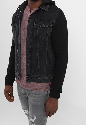 Hombre con chaleco de mezclilla negro con mangas de sudadera negra, camiseta henley color granate y vaqueros grises rotos.