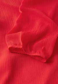 Roter strukturierter Stoff mit fein gerippter Oberfläche und elastischem Rand. Das Material wirkt leicht und fließend und zeigt eine lebendige Farbe.