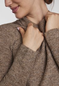 Brown knitted sweater with a high neckline, textured fabric, and ribbed cuffs. Hands clasping the fabric near the neck.