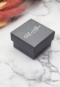 Svart texturerad smyckeslåda med fyrkantig form och lock, med silverfärgad logotyp på toppen mot en marmorbakgrund.