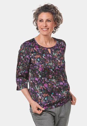 Blumenbluse in Schwarz mit lila, grünen und orangen Mustern. Verfügt über Dreiviertelärmel, einen runden Ausschnitt und ein Schlüsselaugen-Detail.