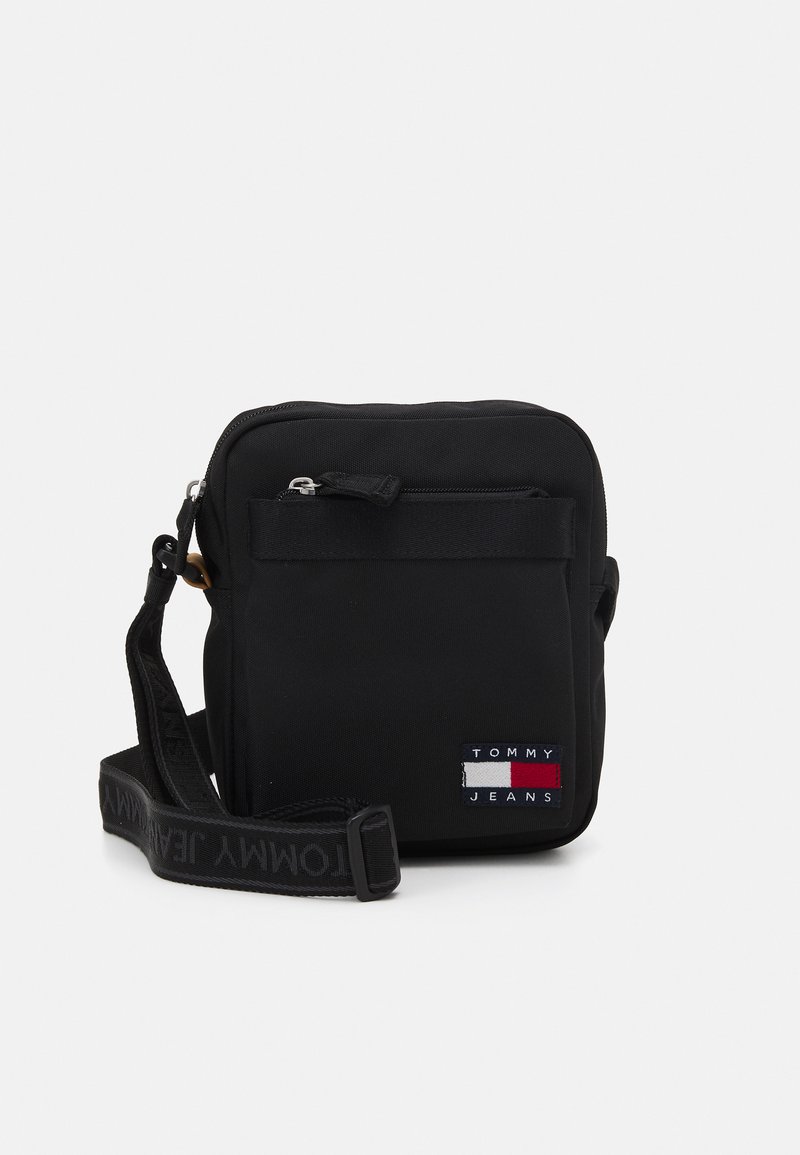 Fekete keresztpántos táska tartós anyagból, cipzáras elülső zsebbel és "TOMMY JEANS" logós pánttal. Téglalap alakú.