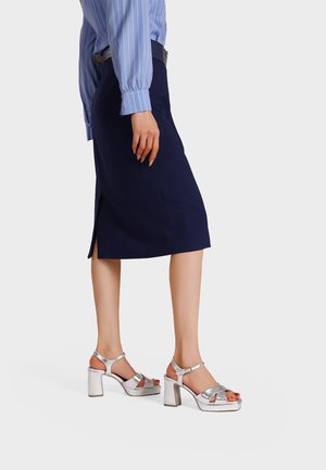 Donna che indossa una gonna blu navy al ginocchio con spacco laterale, camicia a righe blu, cintura nera e sandali argento con tacco alto e punta aperta.