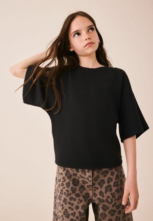 Ung pige med langt brunt hår iført en sort oversized t-shirt og leopardprint bukser, som kigger opad med den ene hånd i håret.