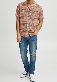 Camisa de manga corta de color marrón con un estampado geométrico; combinada con unos jeans de mezclilla azules y zapatillas blancas con detalles en azul marino.