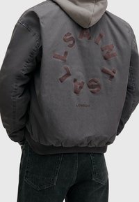 Persona que lleva una chaqueta gris con capucha, con un texto circular que dice "SAINT" repetido y "LONDON" debajo en la parte posterior, combinada con jeans oscuros.