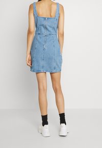 Denimmekko, jossa on neliömäinen selkä, istuva yläosa ja levenevä hame. Vaaleansininen väri, näkyvät sauman yksityiskohdat ja keskiselässä oleva vetoketju.