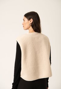Beige fleece vest med rundet nederste kant og en blød tekstur, båret over en langærmet sort top. Bagfra ses det minimalistiske design.
