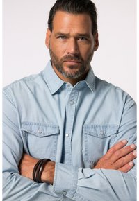 Camicia in denim azzurro chiaro con bottoni a pressione, due tasche sul petto e maniche arrotolate. Presenta una texture morbida e una vestibilità rilassata. Bracciale da polso in pelle scura.