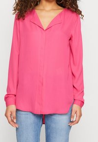 Blouse rose à manches longues avec un col en V, texture douce et fermeture à boutons. Associée à un jean bleu clair, affichant une coupe décontractée.