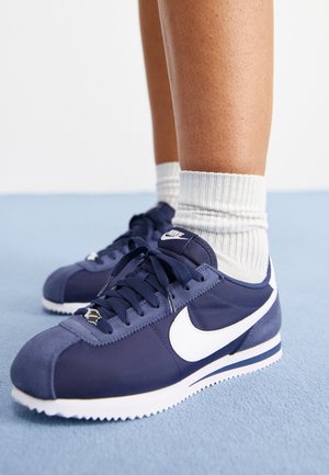 Persona che indossa sneakers Nike blu navy con logo Swoosh bianco e calzini bianchi a coste in piedi su una superficie azzurro chiaro.