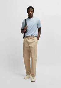 Hellblaues, kurzärmliges T-Shirt, beiger gerader Hosen und cremefarbene Sneaker; Modell hält eine schwarze Umhängetasche.