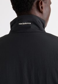 Svart jacka med hög krage, med en vit "new balance" logotyp på baksidan. Tyget verkar lätt och mjukt.
