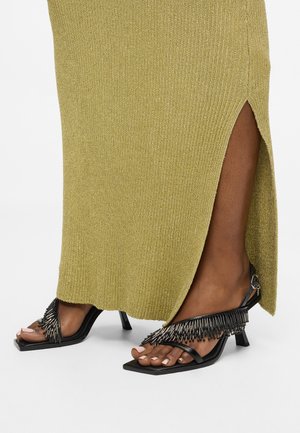 Geribbelde olijfgroene maxi-jurk met een zijspleet, gecombineerd met zwarte block-heeled sandalen versierd met metallic franjes.