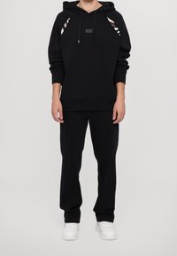 Černá mikina s kapucí s proužkovanými doplňky na ramenou a s nášivkou s logem. Kombinováno s černými kalhotami a bílými teniskami.