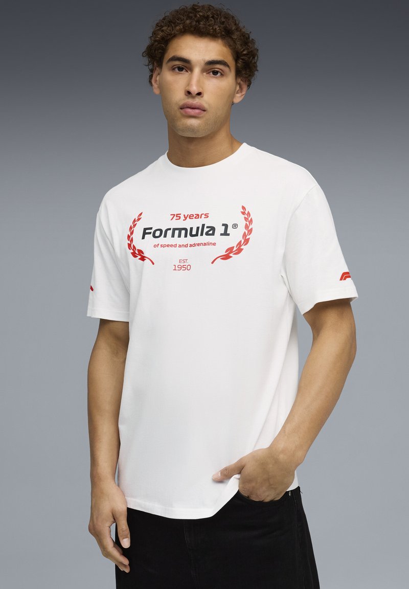 Hvid bomulds t-shirt med rund hals, prydet med rød og sort "Formula 1" grafik og laurbærdesign. Sideetiket på ærmet. Modellen står foran en grå baggrund.