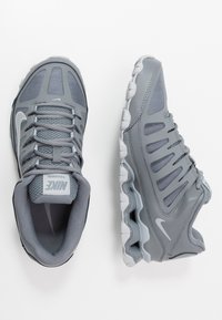 Šedé tenisky Nike s síťovaným svrškem, výrazným systémem šněrování a texturovanými detaily. Podešev obsahuje několik měkkých polštářků pro podporu.