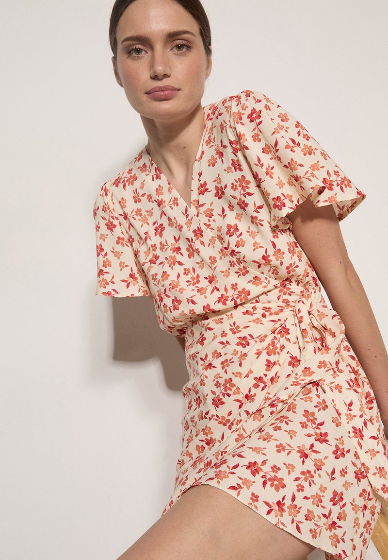 Mujer con un vestido cruzado crema con estampado floral rojo y naranja, mangas cortas de volante, sentada frente a un fondo claro.