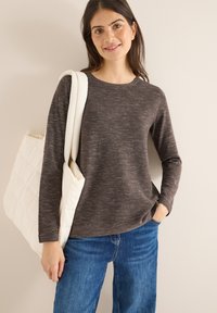 Maglietta lunga a maniche con scollo rotondo di un marrone scuro melangiato, abbinata a jeans blu; tiene una borsa a tracolla bianca trapuntata.