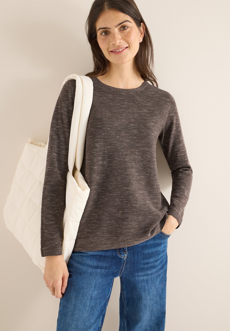 Maglietta lunga a maniche con scollo rotondo di un marrone scuro melangiato, abbinata a jeans blu; tiene una borsa a tracolla bianca trapuntata.