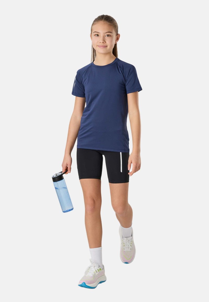 Jeune fille marchant, portant un t-shirt de sport bleu marine, un short noir, des chaussettes blanches, des baskets, et tenant une bouteille d'eau bleue.