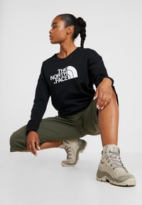 Sweatshirt preto com logo branco, calças verde cortadas e botas de caminhada bege. Tecido suave e design estruturado. Modelo posa numa posição agachada.