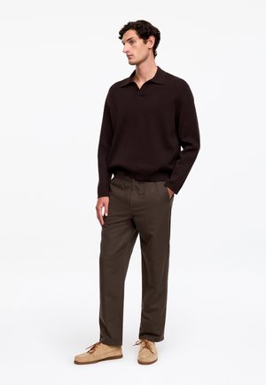 Młody mężczyzna stoi, ubrany w ciemnobrązowy sweter z kołnierzykiem i długimi rękawami, oliwkowe spodnie oraz beżowe zamszowe buty, z rękami w kieszeniach, patrzy na bok.
