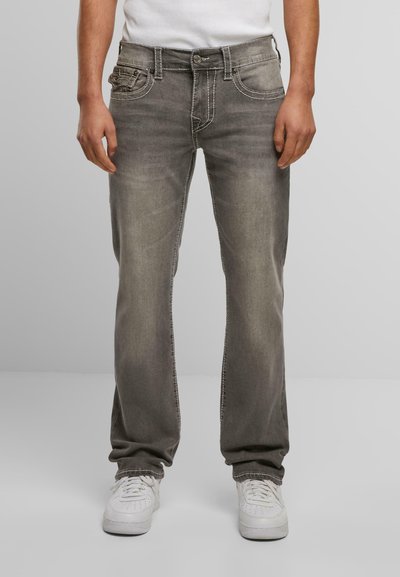 Jean en denim gris avec une jambe droite, décoloration légère et couture contrastante. Dispose de cinq poches et d'une fermeture éclair, porté avec des baskets blanches.