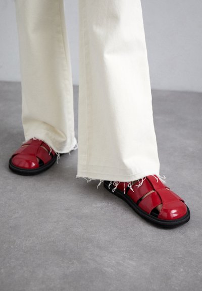 Punaiset nahkakengät, joissa on kudottu muotoilu ja avoin varvas, yhdistettynä vaaleisiin leveälahkeisiin housuihin, joissa on purjeet.