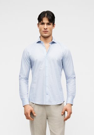 Homme portant une chemise à manches longues bleu clair à motifs et un pantalon beige, debout devant un fond uni clair.