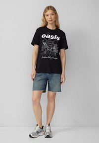 Schwarzes Grafik-T-Shirt mit dem Wort "oasis" und "Definitely Maybe" in Weiß. Kombiniert mit blauen Denim-Shorts und silbernen Turnschuhen.