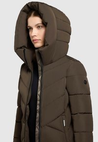 Giacca puffa marrone con un grande cappuccio squadrato, design trapuntato orizzontale e diagonale, zip frontale e una toppa nera con logo sulla manica.