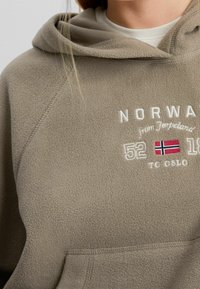 Taupenvärinen fleecetuulipaita etutaskulla. Siinä on kirjailtu teksti "NORWAY from Jørpeland 52 TO OSLO" ja Norjan lippu.
