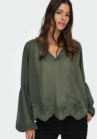 Donna che indossa una blusa trasparente verde scuro con una texture a pois, ricami floreali sull'orlo scallop e maniche lunghe e ampie, abbinata a pantaloni neri.