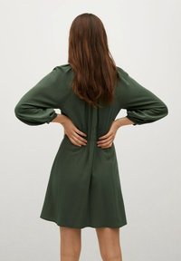 Vihreä mekko, joka on valmistettu sileästä kankaasta; siinä on pitkät hihat, kireä vyötärö ja liehuva hame. Takakatsaus ilman lisätietoja.