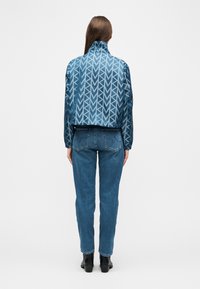 Femme portant une veste bleue à motifs, un jean bleu et des bottines noires, debout de dos sur un fond uni.