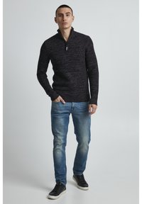 Dunkelgrauer, gestrickter Zip-Hoodie mit einem Stehkragen, kombiniert mit blauen Jeans und schwarzen Sneakers. Mann steht aufrecht vor einem neutralen Hintergrund.