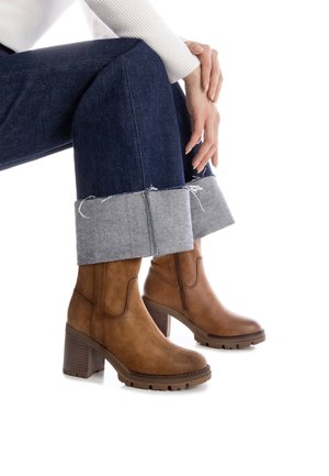 Botas de ante marrón hasta el tobillo con un tacón grueso, que presentan una suela texturizada y paneles elásticos en los laterales, combinadas con unos pantalones recortados de mezclilla oscura.