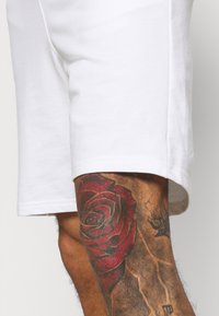 Pantalones cortos de algodón blancos con corte recto, que presentan un dobladillo enrollado. Un gran tatuaje de rosa roja es visible en el muslo superior.