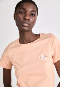 Breskvaste barve bombažna majica s kratkimi rokavi in okroglim izrezom, z belim napisom "Calvin Klein Jeans" na levem prsnem delu. Gladka tekstura.