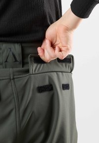 Groene technische broek met klittenbandaccenten en een logo, gekenmerkt door een aansluitende taille en een gladde, lichte stof. Hand die de tailleband vastgrijpt.